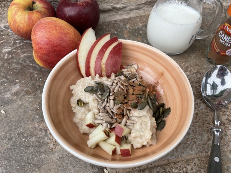 En skål med havregrynsgröt toppad med skivade äpplen, frön och hackade äppelbitar står på en stenyta. Detta nyttiga frukostalternativ omges av hela äpplen, en glaskanna med mjölk, malen kanel och en sked.
