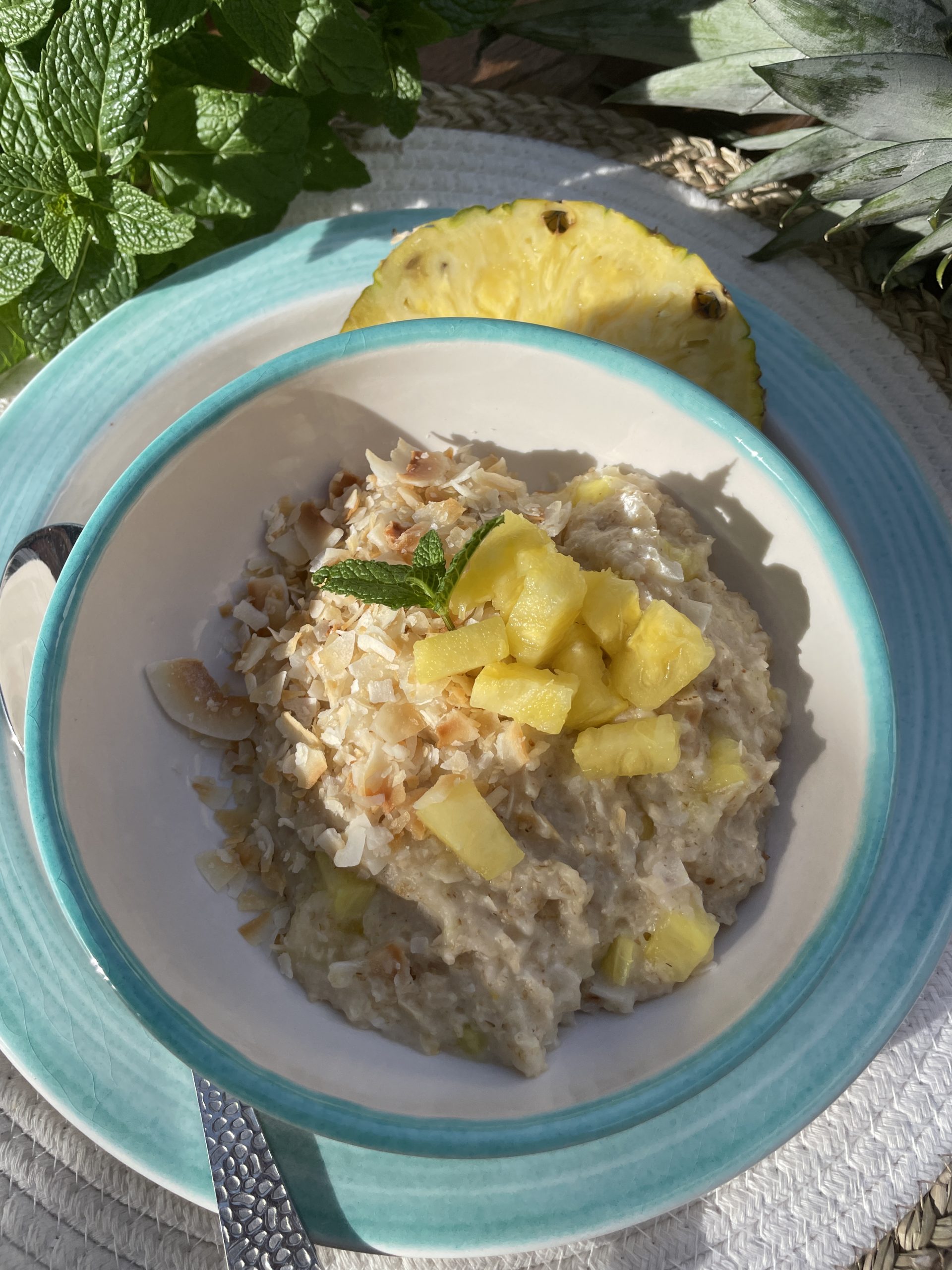 En skål havregrynsgröt toppad med hackad ananas, rostade kokosflingor och en myntakvist, serverad på en turkosrandig tallrik - en inbjudande variant av funktionell mat med livfulla smaker och en skiva ananas i bakgrunden.