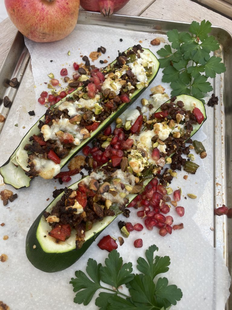 Två zucchinihalvor fyllda med köttfärs, smulad ost, pistagenötter och granatäppelkärnor på en pergamentklädd bricka, garnerad med färsk persilja - ett utsökt exempel på funktionell mat full av smak och näringsämnen.