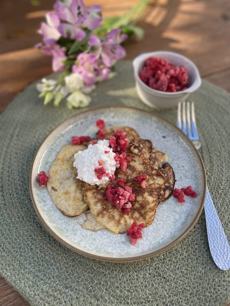 En tallrik med pannkakor toppade med keso och hallonbitar, med extra hallon i en skål bredvid, visar nyttigt och funktionell mat på en grön bordsunderlägg med blommor i bakgrunden.
