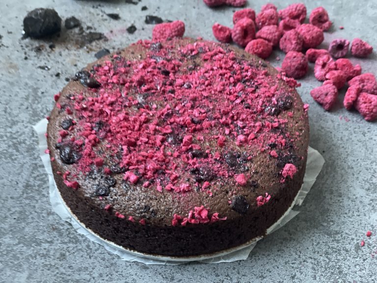 En rund chokladkaka toppad med krossade frystorkade hallon ligger på ett pergamentpapper, med fler hallon utspridda på en grå yta i närheten - ett exempel på hälsosamma recept för den som vill ha nyttigt och gott.