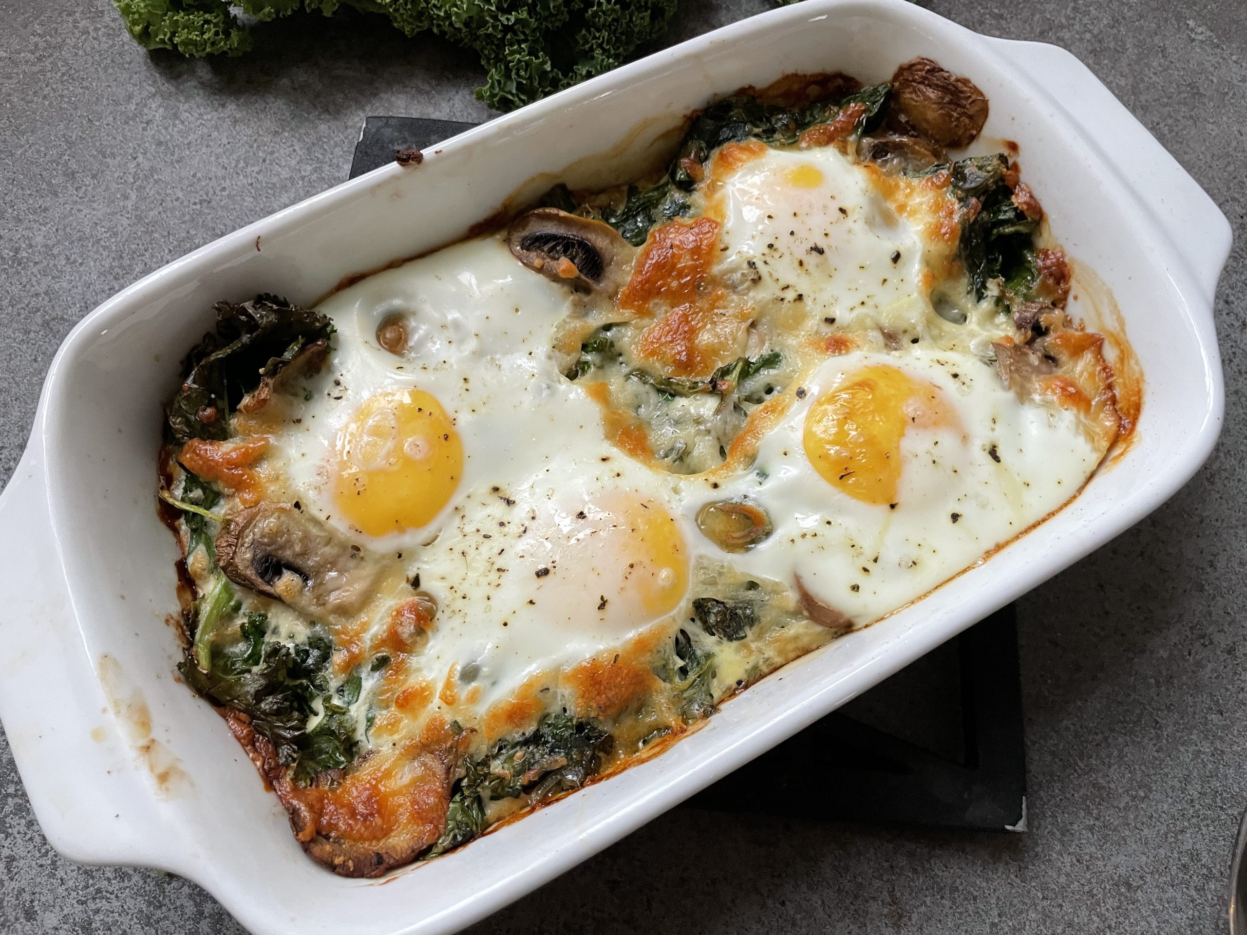 En ugnsbakad ägggryta i en vit skål med tre ägg, svamp, bladgrönsaker och smält ost på toppen -ett nyttigt val för dig som söker hälsosamma recept och funktionell mat.