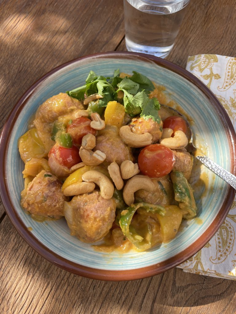 En skål curry med köttbullar, gul paprika, körsbärstomater, cashewnötter och koriander är ett perfekt exempel på hälsosamma recept som serveras på ett träbord med ett glas vatten och en mönstrad servett.