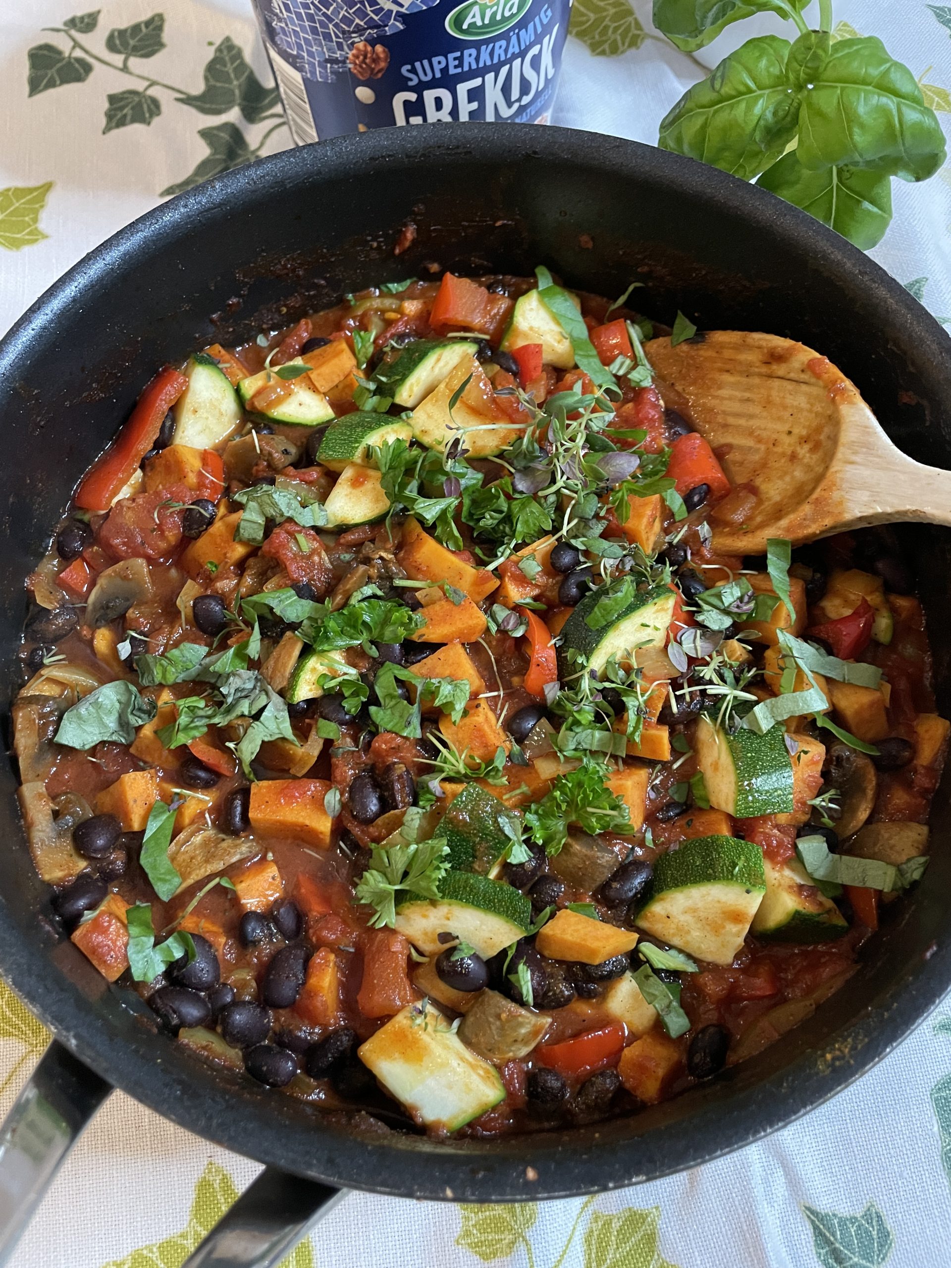 En kastrull fylld med en hälsosamma recept av grönsaker och svart böngryta, inklusive zucchini, morötter, tomater och färska örter, med en träsked vilande inuti.