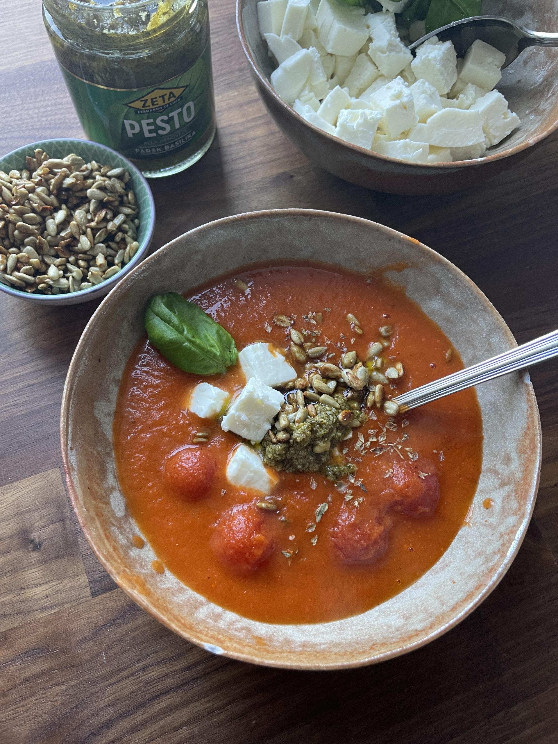 Skål med tomatsoppa toppad med fetaostbitar, pesto, solrosfrön och ett basilikablad - ett läckert exempel på funktionell mat. I omgivningen finns en burk med pesto, en skål med fetaost och en skål med frön.