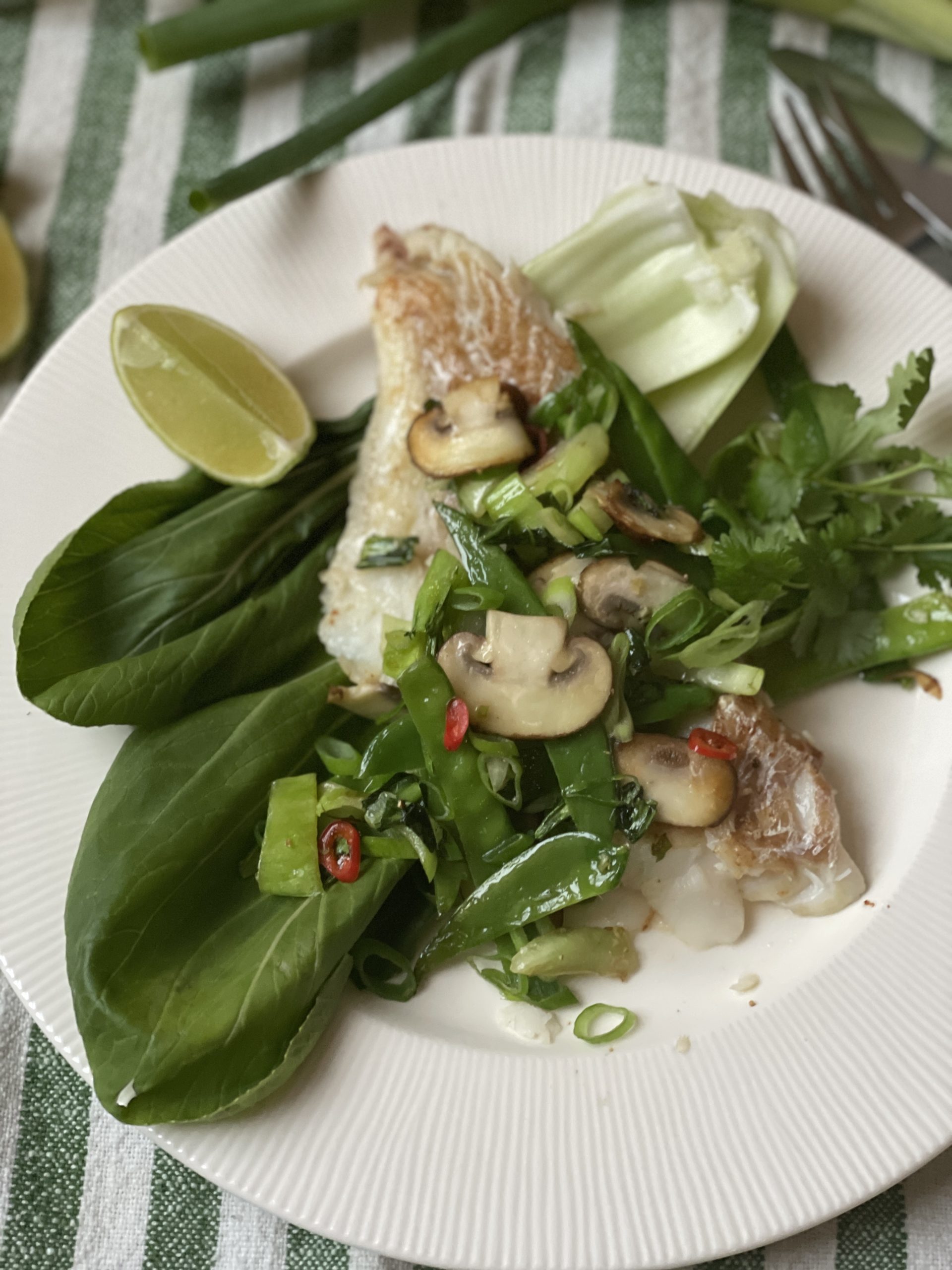 En tallrik med kokt fisk toppad med svamp, gröna grönsaker och skivad röd chili, serverad med bok choy, limeklyfta och färska örter på ett vitt fat - perfekt för dem som söker hälsosamma recept och funktionella livsmedel.