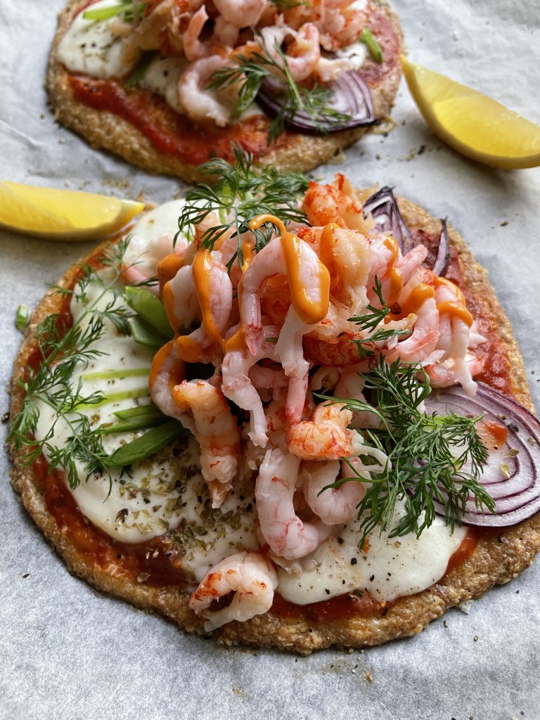 Två skaldjurspizzor toppade med räkor, dill, rödlök, avokado, ost och tomatsås ligger på pergamentpapper med citronklyftor på sidan - perfekt inspiration för hälsosamma recept med funktionella livsmedel.