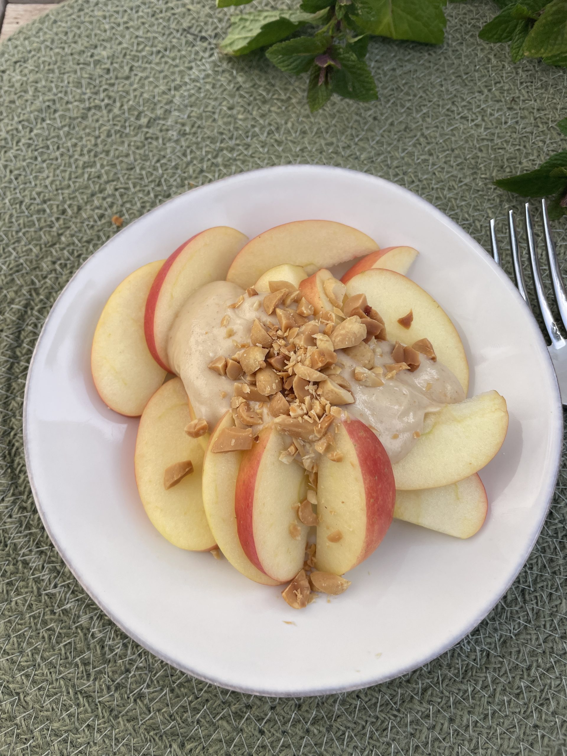 En vit tallrik med skivade äpplen toppade med en krämig röra och hackade jordnötter, placerad på en grön vävd matta med en gaffel och myntablad i närheten - ett inbjudande exempel på hälsosamma recept med funktionell matta.