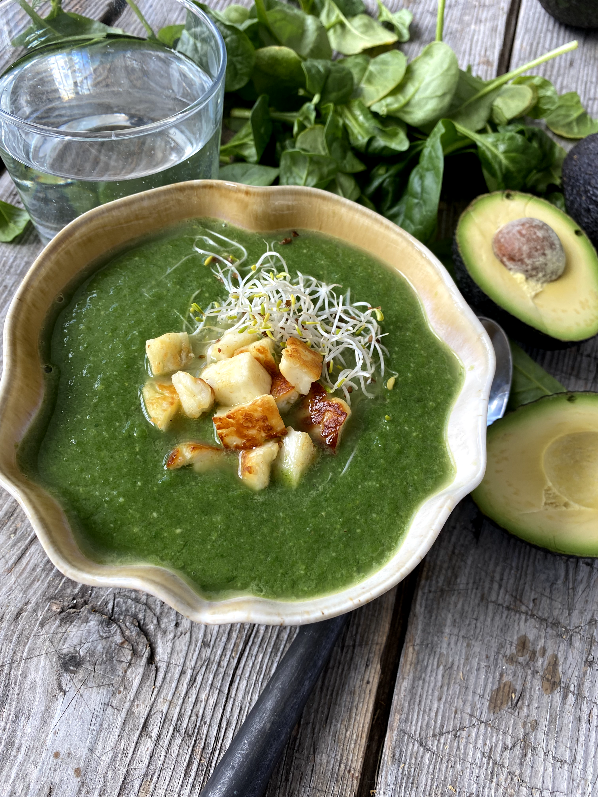 En skål med grön soppa med functional foods toppas med krutonger och groddar och står på ett träbord tillsammans med färsk spenat, en halverad avokado och ett glas vatten.